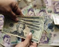 Un hombre sostiene tres billetes de 20 dólares mientras al fondo se observan numerosos billetes colombianos.