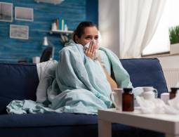 Woman with sickness wrapped in blanket at home ill feeling unwell. Young caucasian adult suffering from temperature fever disease illness flu, taking medical treatment sitting on couch