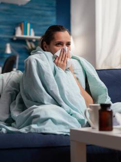 Woman with sickness wrapped in blanket at home ill feeling unwell. Young caucasian adult suffering from temperature fever disease illness flu, taking medical treatment sitting on couch
