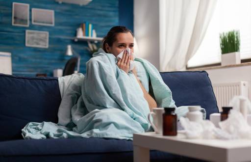 Woman with sickness wrapped in blanket at home ill feeling unwell. Young caucasian adult suffering from temperature fever disease illness flu, taking medical treatment sitting on couch