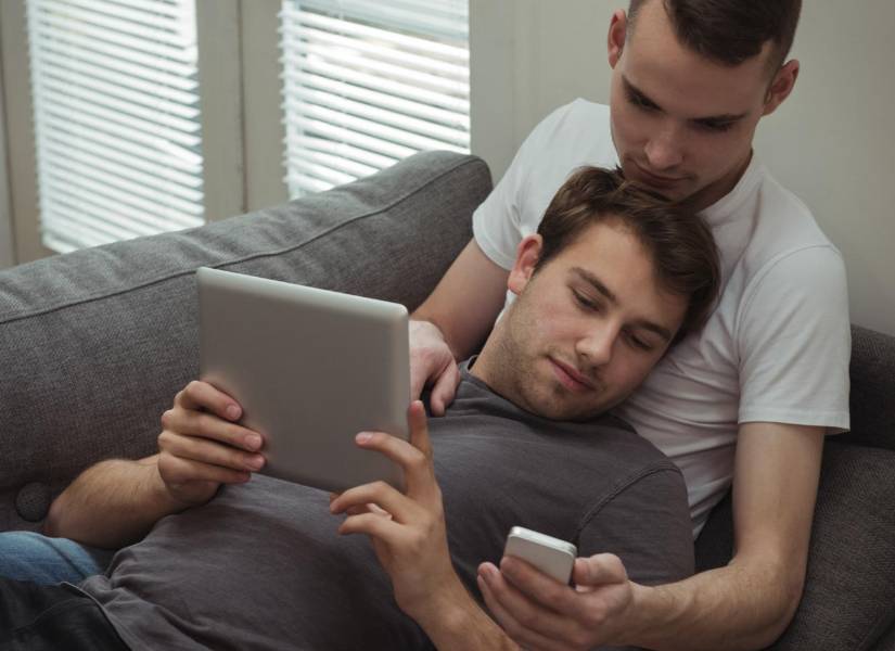 Gay couple using digital tablet and mobile phone while lying on sofa at home